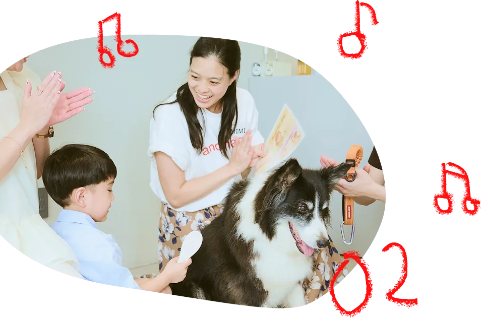 「02」の文字。スタッフたちが手拍子をしながら歌い、子供が犬と隣り合っている写真。